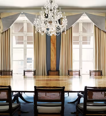 Elegant conference room with a long wooden table surrounded by ornate chairs. Large windows with draped curtains allow natural light to fill the space. A crystal chandelier hangs from the ceiling, adding to the rooms grandeur.