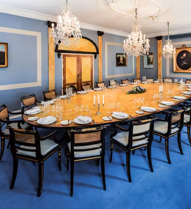 Elegant dining room with a large, oval wooden table set for a formal dinner. Surrounded by black and gold chairs, the room features blue walls, chandeliers, and landscape paintings. A table with a white cloth stands in the corner.
