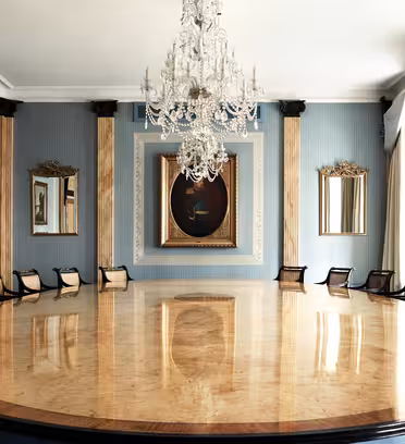 A luxurious meeting room featuring a large, oval wooden table with dark chairs around it. The walls are blue with gold accents and decorated with framed paintings and mirrors. A grand crystal chandelier hangs from the ceiling.