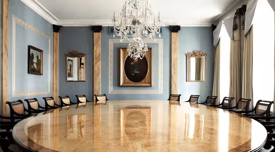 A luxurious meeting room featuring a large, oval wooden table with dark chairs around it. The walls are blue with gold accents and decorated with framed paintings and mirrors. A grand crystal chandelier hangs from the ceiling.
