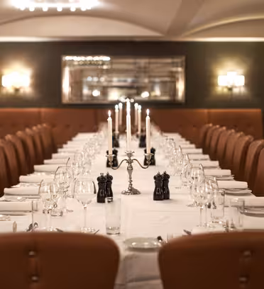 A long, elegant dining table is set with white tablecloths, glassware, and candles in silver holders. Brown leather chairs line both sides. Soft lighting and a large mirror enhance the refined atmosphere.