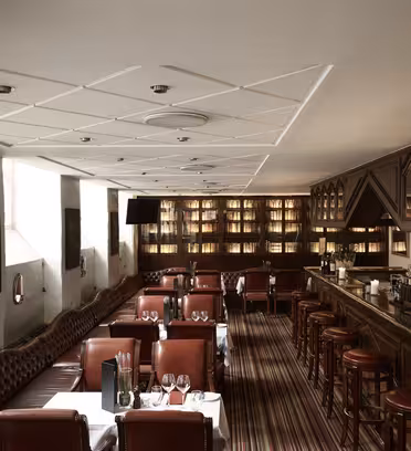 A stylish restaurant interior featuring leather seating, a long bar with stools, neatly set tables with white tablecloths, and shelves filled with bottles. The lighting is warm, and the atmosphere is cozy and inviting.