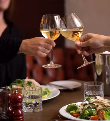 Two people clink glasses of white wine at a warmly lit restaurant table. Plates with salad and meat are on the table, along with a candle, a pepper grinder, and a wine cooler holding a bottle.