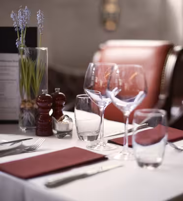 A table set for two in a restaurant with red napkins, wine glasses, and cutlery. A menu and a vase with purple flowers are placed at the center. Red cushioned chairs are in the background.