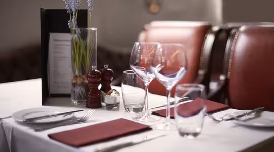 A table set for two in a restaurant with red napkins, wine glasses, and cutlery. A menu and a vase with purple flowers are placed at the center. Red cushioned chairs are in the background.