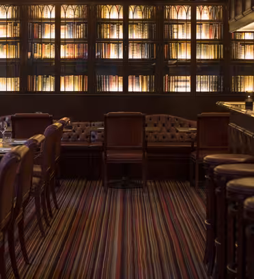A cozy, dimly lit bar with leather stools and wooden tables set for dining. The walls are lined with tall bookshelves filled with books, creating a warm and inviting atmosphere. The floor has a colorful striped carpet.