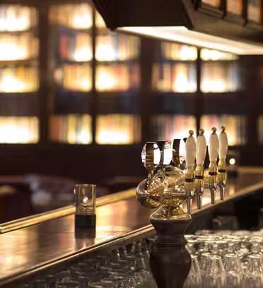 Cozy bar interior with a polished wooden counter, featuring beer taps and neatly arranged glasses. Warm lighting highlights a backdrop of shelves filled with books, adding a sophisticated ambiance.
