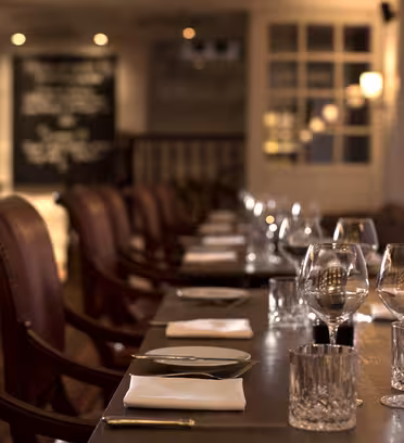 A dimly lit, elegant restaurant interior showing a side view of a wooden dining table set with white napkins, glassware, and cutlery. The background features soft lighting and a blurred chalkboard menu.