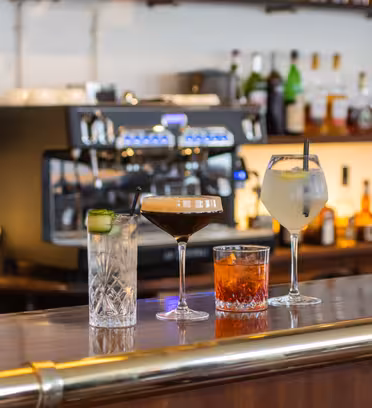 Four cocktails are lined up on a wooden bar in front of a coffee machine and bottles. The drinks include a clear drink with cucumber, a dark drink with foam, an orange drink in a rocks glass, and a light drink in a wine glass with a straw.
