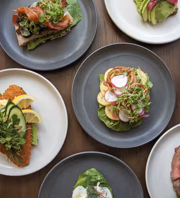 Assorted plated dishes on a wooden table. Includes open-faced sandwiches with toppings like avocado, eggs, bacon, leafy greens, and shrimp. Plates are a mix of gray and white, with ingredients artistically arranged.