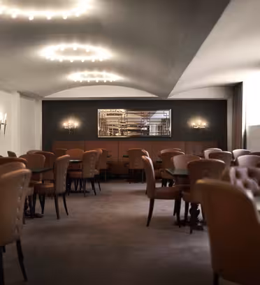A dimly lit, elegant dining room featuring leather chairs and round tables. Warm lighting with decorative wall sconces enhances the cozy atmosphere. A large mirror hangs on the back wall, reflecting the rooms details.