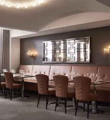 A modern dining area with a long, brown leather bench and matching chairs around tables set with empty wine glasses. A large, decorative mirror and wall sconces are mounted on the dark accent wall. Ceiling lights illuminate the space.
