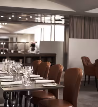 Elegant restaurant interior with neatly set tables featuring white napkins and wine glasses. Soft lighting and brown leather chairs create a warm ambiance. Background shows a partition with more seating, under a ceiling with recessed lighting.