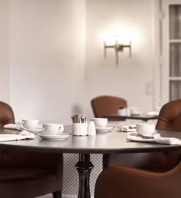 A round table in a softly lit café is surrounded by plush brown chairs. On the table are white cups and saucers, white napkins, and a caddy with sugar packets. A soft glow emanates from a wall sconce in the background.