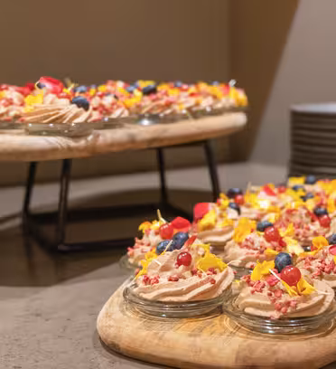 Small dessert cups filled with cream, topped with berries and edible flowers, displayed on wooden trays. Plates are stacked in the background.