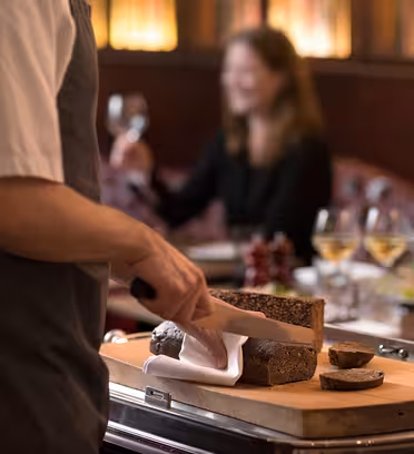 A person wearing an apron slices brown bread on a wooden board with a large knife in a restaurant setting. In the background, a woman holds a glass of white wine with more glasses on the table. The atmosphere is warm and inviting.