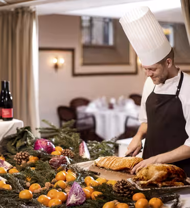 A chef in a tall white hat and brown apron slices meat on a table adorned with greenery, tangerines, pinecones, and red cabbage. Wine bottles are displayed nearby. The setting is an elegant dining room with a table visible in the background.