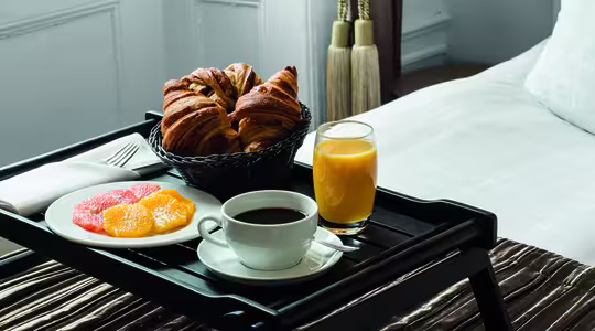 Breakfast tray on a bed with a cup of coffee, a plate of sliced citrus fruits, a basket of croissants, and a glass of orange juice. The setting is elegant and cozy, with soft lighting and a pillow in the background.