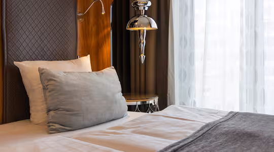 A neatly made bed with white linens and a gray pillow is beside a wooden panel with a silver lamp. Sheer curtains allow soft light to enter the room, creating a tranquil ambiance.